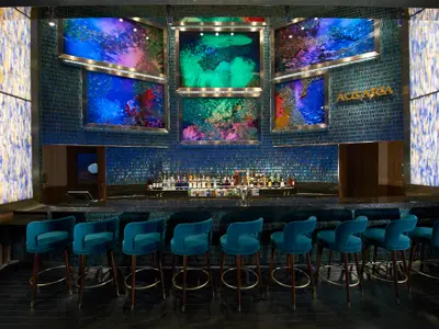 Übersichtsbild der modernen AQUARIA Bar auf der Carnival Celebration. Die Bar ist mit blauen Mosaikfliesen dekoriert und verfügt über mehrere große, beleuchtete Kunstinstallationen mit Unterwassermotiven. Eine Reihe türkisfarbener Samt-Barhocker steht vor einer gut gefüllten Bar mit Spirituosenflaschen und Gläsern.