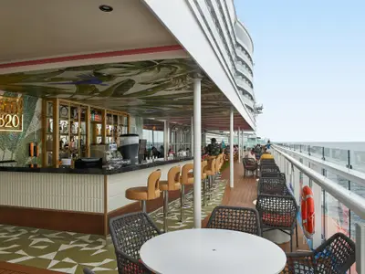 Blick auf eine moderne Außenbar auf dem Deck der Carnival Celebration. Eine glitzernde Mosaikwand, ein goldenes Logo und eine tropisch bemalte Decke prägen den Bereich. An der Bar sitzen Gäste, weitere entspannen auf der Terrasse mit Meerblick.