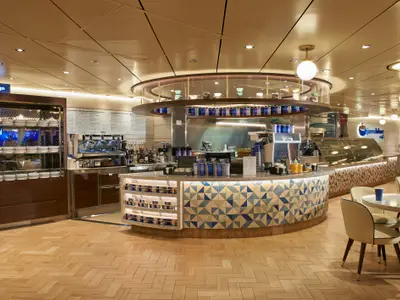 Innenansicht des Javablue Cafés auf der Carnival Celebration mit einer geschwungenen Theke mit blauen Fliesen und Kaffeemaschinen. Rechts sind Tische mit Stühlen und links ein Grab & Go Bereich zu sehen.