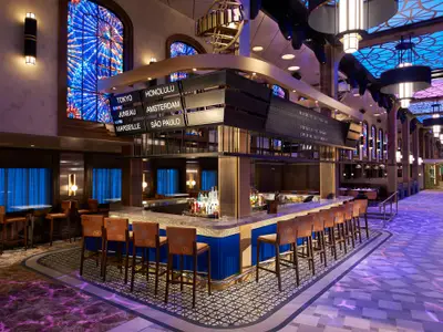 Eine elegant beleuchtete Bar mit einem großen Display, das Reiseziele wie Tokyo und Honolulu anzeigt. Hocker säumen die Theke, Tische stehen im Hintergrund. Das Ambiente ist modern und einladend mit blauen und goldenen Akzenten. Blaue Vorhänge sind in den Fenstern zu sehen.