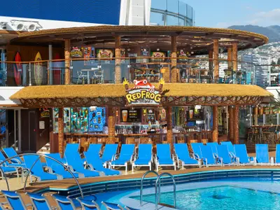 Die RedFrog Tiki Bar auf der Carnival Celebration: Strohdach, bunte Surfbretter, blaue Liegestühle am Pool. Im Hintergrund eine Küstenstadt.