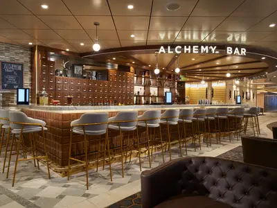 Innenansicht der Alchemy Bar mit langer Theke, Rückenwand voller Holzschubladen, grauen Barhockern mit goldenen Füßen und Deckenbeleuchtung. Rechts dunkle Ledersofas.