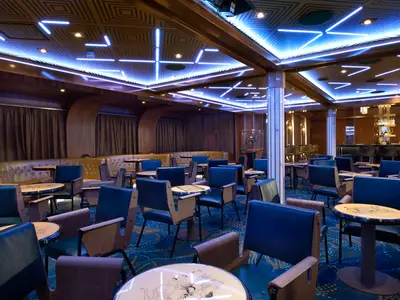 Elegante Bar-Lounge der Carnival Celebration. Blaue Neonlichter an der Decke, dunkles Holz, cognacfarbene Bänke, blaue Stühle und Tische. Im Hintergrund eine beleuchtete Bar. Keine Personen.