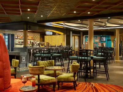 Eine einladende Bar-Lounge an Bord der Carnival Celebration mit gemusterten Fliesen, bequemen Hockern und Sesseln, hohen Tischen und einem beleuchteten Bar-Tresen mit Flaschen und Gläsern. Im Vordergrund sind zwei Cocktails zu sehen. Das Interieur ist im kubanischen Stil gehalten, erkennbar am Neon-Schild 'Havana Bar'.