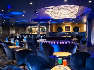 Luxuriöse, tiefblaue Bar-Lounge auf der Carnival Celebration. Elegante Samtsessel, Tische und ein Flügel sind zu sehen. Ein großer Kristallleuchter und eine beleuchtete Bar schaffen eine stimmungsvolle Abendatmosphäre. Keine Personen zu sehen.
