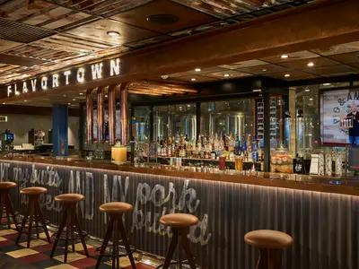 Eine moderne Bar mit dem leuchtenden Schriftzug 'FLAVORTOWN' an der Decke. Die Theke ist mit braunen Barhockern bestückt und zeigt eine große Auswahl an Spirituosen. Im Hintergrund ist ein Fernsehbildschirm mit Guy Fieri zu sehen.