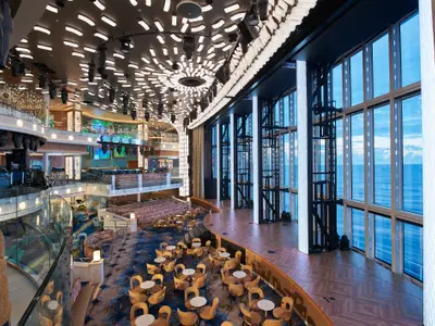 Elegante, mehrstufige Lounge auf der Carnival Celebration mit moderner Deckenbeleuchtung und großen Fenstern zum Meer. Viele Sitzbereiche, Bühne und Bar sind sichtbar.