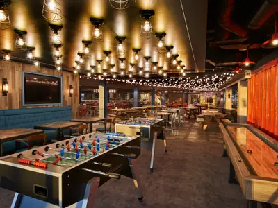 Geräumiger Innenraum der Carnival Celebration mit mehreren Tischen, Bänken, zwei Tischfußballtischen und einem Shuffleboard-Tisch. Die Decke ist mit Industrieleuchten und Lichterketten geschmückt, die Wände sind teilweise holzvertäfelt und eine Seite ist mit leuchtend rotem Wellblech verkleidet. Der Raum ist hell beleuchtet und wirkt einladend.