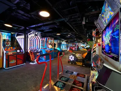 Eine moderne Spielhalle auf der Carnival Celebration mit einer Vielzahl von leuchtenden Arcade-Automaten, darunter Basketballspiele, Greifautomaten und ein Tanzspiel mit roten Haltegriffen. Die Decke ist dunkel mit sichtbaren Rohren und Beleuchtung. Keine Personen sind zu sehen.