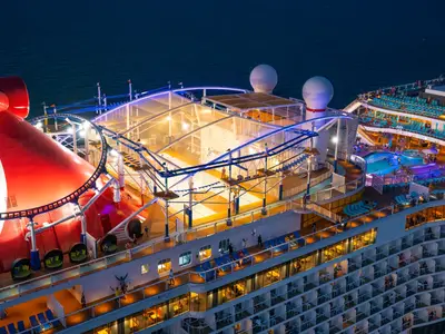 Luftaufnahme des beleuchteten Oberdecks der Carnival Celebration bei Nacht. Man sieht eine Achterbahn um den Schornstein, einen Seilgarten über einem Sportplatz und einen Poolbereich mit zahlreichen Liegestühlen.