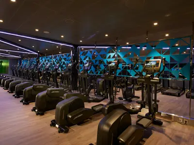 Reihe moderner schwarzer Ellipsentrainer der Marke Life Fitness in einem beleuchteten Fitnessstudio auf der Carnival Celebration. Die Wände sind mit blauen geometrischen Mustern verziert, der Boden ist aus hellem Holz. Im Hintergrund sind Spiegel und weitere Geräte sichtbar.