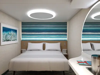Eine modern gestaltete Kabine der Carnival Celebration mit einem Doppelbett und weißer Bettwäsche. Die Wand hinter dem Bett zeigt ein blau-türkises Streifenmuster, die linke Wand ein helles Korallenriff-Bild. Eine runde Deckenleuchte spendet warmes Licht.
