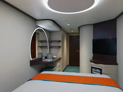 Eine moderne Kabine der Carnival Celebration mit einem Bett im Vordergrund, einem beleuchteten ovalen Spiegel über einem eleganten Schminktisch, Regalen und einem Flur mit einer Tür im Hintergrund. Rechts ein TV und ein Mini-Kühlschrank. Der Boden hat ein türkises Muster.