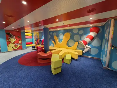 Farbenfroher Kinderbereich an Bord im Dr. Seuss-Stil mit Sam-I-Am und Kater mit Hut. Bunte, weiche Sitzmöbel in Gelb, Rot und Grün praegen den Raum.