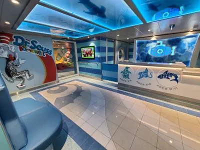 Empfangsbereich eines Kinderclubs auf dem Schiff. Dr. Seuss 'Cat in the Hat' an der Wand. Empfang mit Logos für Altersgruppen 2-5, 6-8 und 9-11. Maritime Farbgebung.