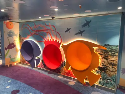 Farbenfroher Kinderbereich auf der Carnival Celebration. Die Wand ist mit einem Unterwasser-Wandbild bemalt, das Quallen, Fische, Krabben und Robben zeigt. Drei große, bunte, leuchtende Röhren, die wie Fische geformt sind, ragen aus der Wand heraus und dienen als Kletterobjekte. Der Teppich ist lila.