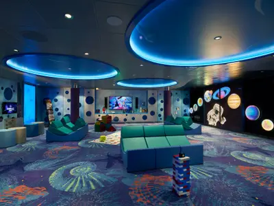Ein modern eingerichteter Kinder- und Jugendbereich auf der Carnival Celebration. Die Szene zeigt bequeme, modulare Sofas, mehrere Bildschirme für Videospiele, eine große Projektionswand mit Weltraummotiven und Planeten sowie einen Meeres- oder Weltraumteppich in Blau- und Grüntönen. Die Decke hat futuristische Lichtinstallationen. Der Raum ist kinderfreundlich und verspielt gestaltet.