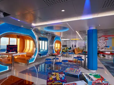 Moderne und farbenfrohe Lounge auf der Carnival Celebration mit verschiedenen Sitzbereichen, großen Fenstern mit Meerblick, Gesellschaftsspielen auf dem Boden und Tischen. Die Einrichtung umfasst leuchtende Blautöne und warme Orangetöne.