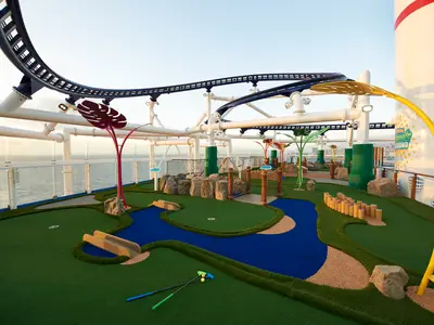 Bunter Minigolfplatz auf dem Deck der Carnival Celebration. Grüne Bahnen, ein blauer Fluss, bunte Deko-Pflanzen, und im Hintergrund das Meer sowie eine Achterbahn.