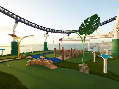 Farbenfroher Minigolfplatz auf dem Oberdeck der Carnival Celebration. Künstliches Gras, bunte Hindernisse, Meer und Stadt im Hintergrund, Achterbahnstrecke oben.