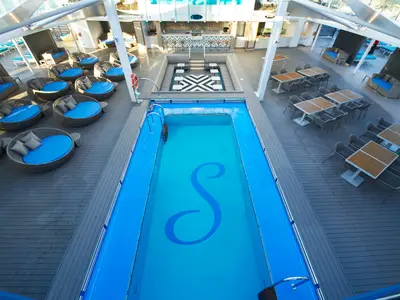 Vogelperspektive auf das Sonnendeck der Carnival Celebration. Ein langer, blauer Pool mit 'S'-Logo, umgeben von runden Lounge-Betten mit hellblauen Polstern und mehreren Esstischen mit Stühlen.