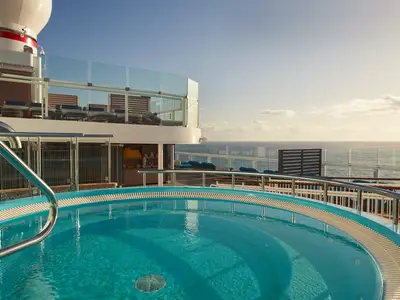 Ein runder Whirlpool mit türkisfarbenem Wasser auf dem Oberdeck der Carnival Celebration. Im Hintergrund sind Liegestühle, eine Glasbalustrade und das weite Meer unter einem hellen Himmel zu sehen.