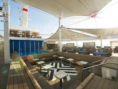 Helles Sonnendeck auf der Carnival Celebration. Blauer Pool links. Großer Lounge-Bereich mit Rattan-Sesseln, Sofas und Tischen unter weißen Sonnensegeln spendet Schatten. Ohne Personen.