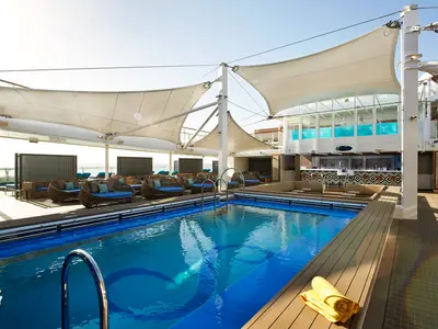 Sonniges Pooldeck der Carnival Celebration. Blauer Swimmingpool umgeben von Holzdecks und Rattan-Lounge-Möbeln mit blauen Kissen. Eine moderne Bar mit gemustertem Tresen und hohe Sonnensegel spenden Schatten. Das Meer ist in der Ferne sichtbar.