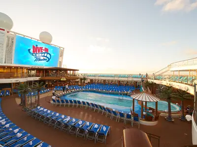 Weitwinkelansicht des Pooldecks auf der Carnival Celebration bei Sonnenuntergang. Ein großer Pool ist von zahlreichen blauen Liegestühlen umgeben, viele davon mit weißen Handtüchern. Im Hintergrund ist eine riesige LED-Leinwand mit der Aufschrift "DIVE-IN Movies" zu sehen. Mehrere Menschen sind auf dem Deck unterwegs. Rechts gibt es einen Whirlpool mit Sonnenschirm.