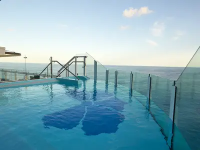 Ein Infinity-Pool auf der Carnival Celebration bei Tag. Das klare, blaue Wasser des Pools geht nahtlos in den weiten Ozean über. Glaswände und Geländer trennen den Pool vom Meer. Ein leicht bewölkter Himmel spannt sich darüber. Ohne Personen.