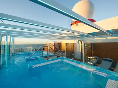 Moderner Poolbereich auf der Carnival Celebration bei Sonnenuntergang. Zwei Pools und ein Whirlpool mit blauen LED-Streifen unter einer Pergola. Meerblick. Liegestühle am Beckenrand.
