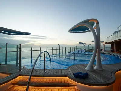 Blick auf das moderne Pooldeck der Carnival Celebration bei Sonnenaufgang. Infinity-Pool, Holzdeck, stilvolle weiße Strukturen, Meerblick. Blaue Handtücher bereitliegend.