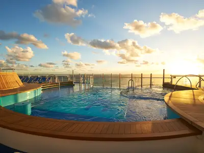 Blick auf das leere Pooldeck der Carnival Celebration bei Sonnenuntergang. Holzdecks umgeben einen Pool mit Meerblick, während die Sonne am Horizont untergeht und den Himmel orange färbt.