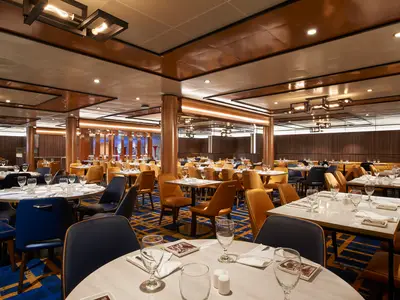 Ein geräumiges Restaurant der Carnival Celebration mit runden und eckigen Tischen, gedeckt mit Gläsern und Servietten. Dunkelblaue und senffarbene Stühle stehen auf einem gemusterten Teppich. Warme Holzpaneele und moderne Beleuchtung prägen den Raum.