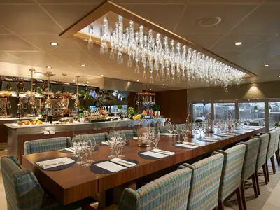 Stilvolles Restaurant auf der Carnival Celebration mit langer Holzetafel und Buffet. Eine kunstvolle Lichtinstallation aus Trinkgläsern schmückt die Decke.