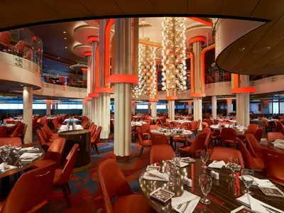 Innenansicht eines mehrstöckigen Restaurants der Carnival Celebration mit zahlreichen Tischen und roten Stühlen. Auffällige Säulen und glänzende Kronleuchter prägen das elegante Ambiente.