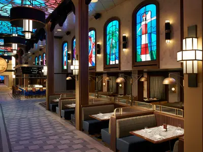 Ein stilvolles Restaurant auf der Carnival Celebration mit farbigen Buntglasfenstern, bequemen Sitzecken, einer langen Bar und moderner Beleuchtung. Der Boden hat ein geometrisches Fliesenmuster.