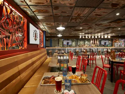 Innenansicht eines modernen Restaurants an Bord der Carnival Celebration. Rote Stühle, Holzbänke und Tische mit Speisen und Getränken. Im Hintergrund eine Bar mit beleuchtetem 'FLAVORTOWN'-Schriftzug.