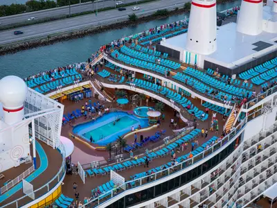 Blick von oben auf die lebhaften oberen Decks der Carnival Celebration mit zwei Pools, unzaehligen türkisen Sonnenliegen und vielen entspannten Passagieren.