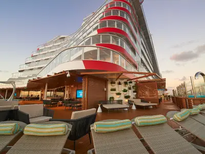 Ein modernes Sonnendeck der Carnival Celebration mit bequemen Liegen, Sitzbereichen, einer Bar und einer künstlerischen Fassade bei Sonnenuntergang.