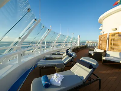 Ein leeres Sonnendeck auf der Carnival Celebration mit Reihen von Liegestühlen und gemütlichen Sitzgruppen aus Rattan. Eine transparente Glaswand bietet Schutz vor Wind und freie Sicht auf das weite, blaue Meer und den Horizont. Der Himmel ist wolkenlos.