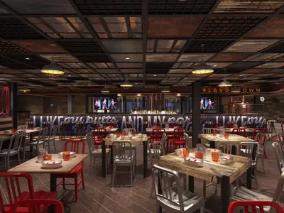 Ein lebhaftes Spezialitätenrestaurant im Industriestil auf der Carnival Celebration. Es gibt Holztische, rote und silberne Stühle, eine lange Bar mit Bildschirmen und eine Decke aus Metallplatten. Keine Personen zu sehen.
