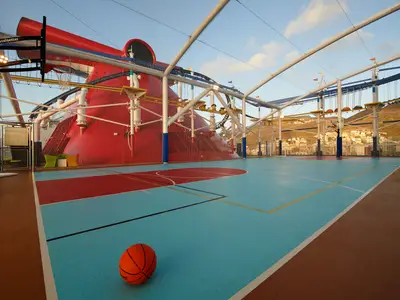 Multifunktionaler Sportplatz auf einem Deck der Carnival Celebration mit Basketballkorb, blau-rotem Boden und einem Basketball im Vordergrund. Im Hintergrund sind ein Hochseilgarten, eine rote Röhrenrutsche und Berge mit einer Stadt zu sehen. Der Himmel ist hellblau mit leichten Wolken.