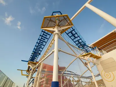 Blick von unten auf einen gelb-blauen Kletterpark mit Seilleitern und Netzen auf der Carnival Celebration. Rote Wasserrutsche und blauer Himmel sichtbar.