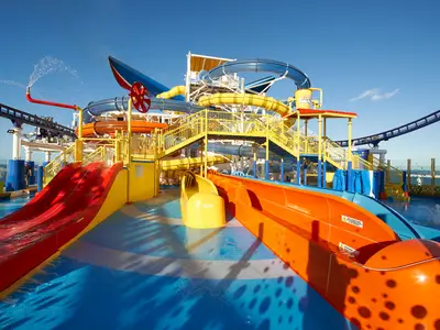 Eine lebhafte Wasserrutschenanlage auf dem Oberdeck der Carnival Celebration mit roten, gelben, orangen und blauen Rutschen. Ein blauer Himmel ist im Hintergrund zu sehen. Wasser spritzt aus einem Rohr und läuft durch die Rutschen.
