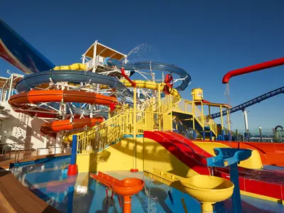 Lebhafter Wasserpark auf der Carnival Celebration. Mehrere bunte Wasserrutschen, darunter blaue, orange und gelbe Röhren, winden sich um eine große gelbe Spielstruktur mit Treppen und Wasserkanonen. Blaues Wasser spritzt aus verschiedenen Elementen in die flachen Pools. Der Himmel ist strahlend blau.