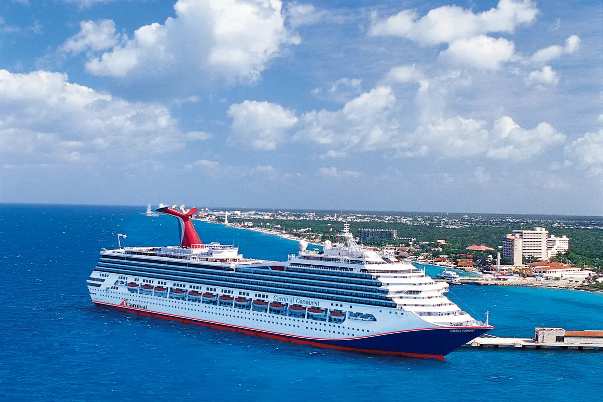 Eine weiß-blaue Carnival Conquest mit rotem Schornstein ankert im leuchtend blauen Meer vor einer belebten Küstenstadt mit Palmen, Strand und weißen Gebäuden unter einem blauen Himmel.