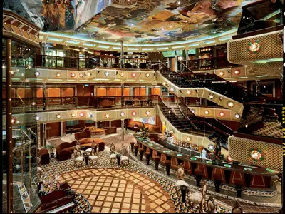 Luxuriöses, mehrgeschossiges Atrium der Carnival Conquest mit großer Bar, eleganten Sitzgruppen und geschwungenen Treppen. Die farbenfroh bemalte Decke und funkelnde Lichter prägen das Ambiente. Keine Personen.