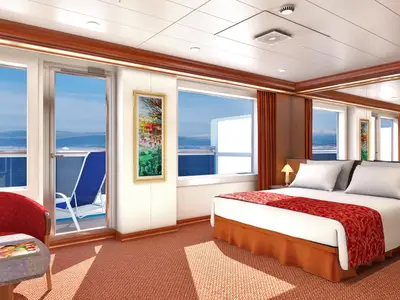 Geräumige Suite auf der Carnival Conquest mit Doppelbett, rotem Teppich, einem roten Sessel und Tisch, sowie einem Balkon mit Blick auf das Meer und schneebedeckte Berge in der Ferne.