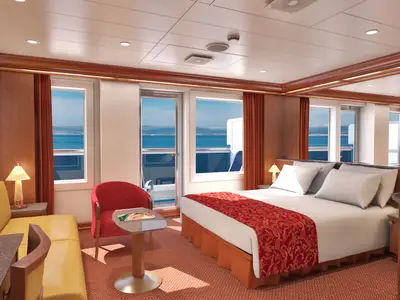 Geräumige Balkonkabine auf der Carnival Conquest mit hellem Doppelbett, rot gemusterter Tagesdecke, gelbem Sofa und Stuhl, sowie einem Balkon mit Meerblick und Küstenlinie im Hintergrund.